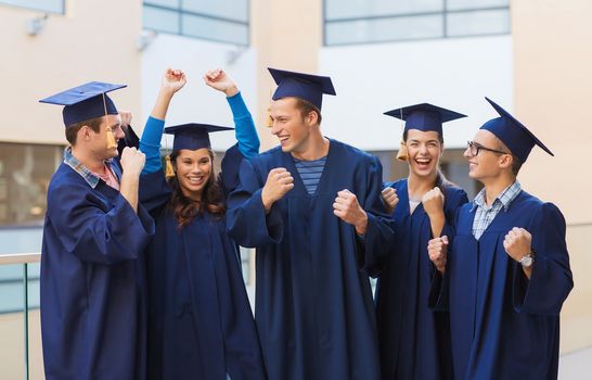 education, graduation and people concept - group of smiling students in mortarboards and gowns making triumph gesture outdoors