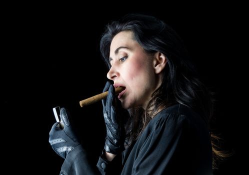 Brunette woman in black dress and black gloves standing in front of a black backdrop, getting ready to light a cigar.
