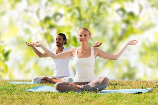 sport, fitness, yoga and people concept - smiling couple meditating and sitting on mats over green tree leaves background