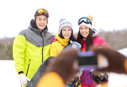 winter sport, leisure, friendship, technology and people concept - happy friends with snowboards and smartphone taking picture