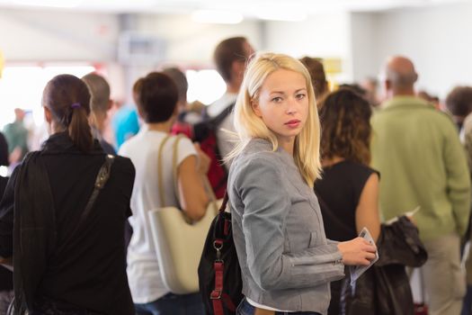 Young blond caucsian woman waiting in line with plain ticket in her hands. Lady standing in a long queue to board a plane.