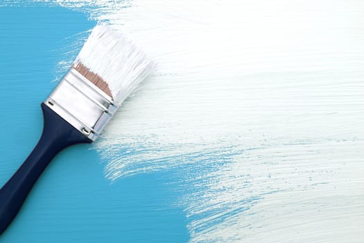Paintbrush with white paint painting over light blue colour on a pine board