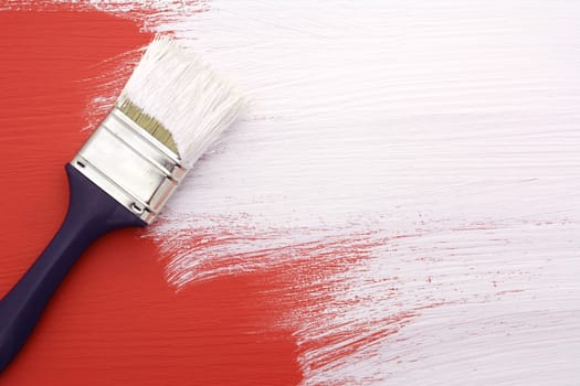 Paintbrush with white paint painting over red colour on a pine board