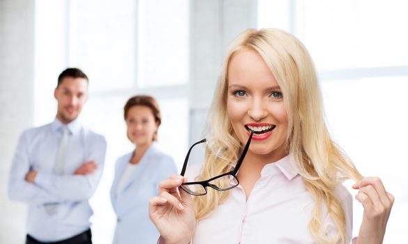 business, teamwork and people concept - smiling businesswoman, student or secretary with eyeglasses over office and group of colleagues background