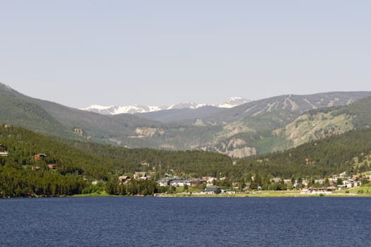 Waters of Barker Meadow Reservoir are foreground to town of Nederland in Colorado.  Summer image shows unmelted snow in Indian Peaks Wilderness behind town.  Evergreens cover slopes of hills.  Horizontal image with copy space in pale blue sky above rural gateway  to nearby Rocky Mountain National Park and Roosevelt Forest.  