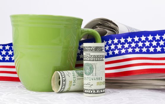 American currency placed in front of newspaper, flag design ribbon, and green mug on white linen