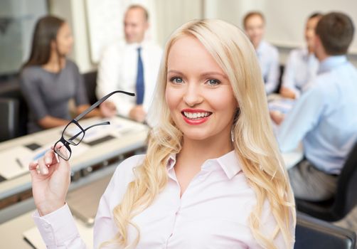 business, teamwork and people concept - smiling businesswoman, student or secretary with eyeglasses over office and group of colleagues background