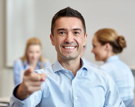 business, people, gesture and teamwork concept - smiling businessman pointing finger to you with group of businesspeople meeting in office