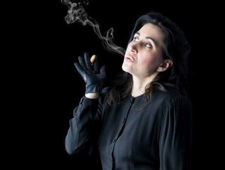 Brunette woman in black dress and black gloves standing in front of a black backdrop, holding a cigar in one hand and blowing smoke.