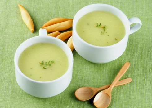 Delicious Cream Asparagus Soup in Two White Soup Cups with Bread Sticks and Wooden Spoons closeup on Green Napkin