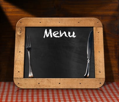 Old blackboard with wooden frame, silver cutlery and text Menu on a table with a checkered tablecloth and wooden wall. Template for recipes or food menu