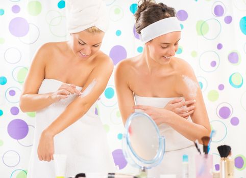 Two young cute woman preparing to start their day. They applying body lotion.