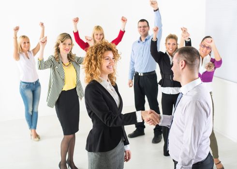 Two successful business people shaking hands at the meeting. Group their colleague celebrating in the background.