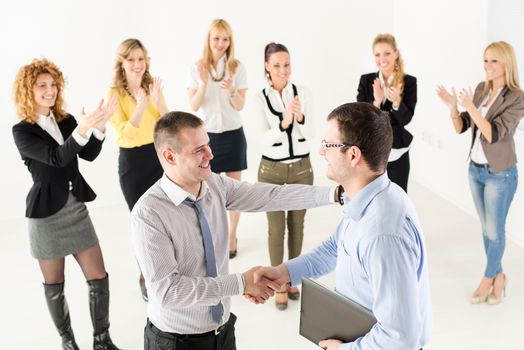 Two successful businessmen shaking hands at the meeting. Group of their colleague celebrating in the background.