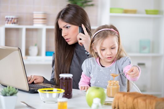 Overworked Business Woman and her little daughter in the morning. Mother read mail and make phone calls before going to work. Daughter smearing peanut butter on bread.