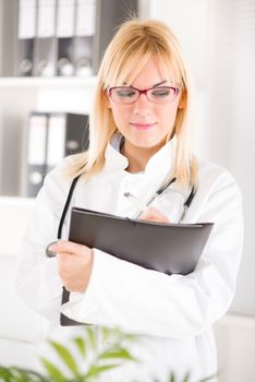 Portrait of a young woman doctor with stethoscope standing in her office and holding clipboard.
