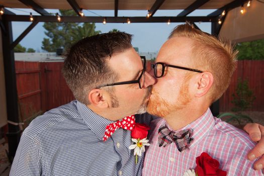 Two men with eyeglasses kiss at outdoor marriage ceremony