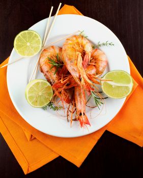Delicious Roasted Shrimps in  with Lime and Rosemary on White Plate with Orange Napkin closeup on Dark Wooden background. Top View