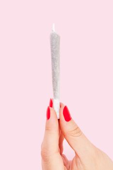 Female hand with red fingernails holding cannabis joint isolated on pink background. Female marijuana abuse. Woman holding joint.