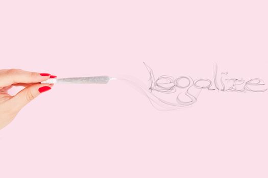 Female hand with red fingernails holding cannabis joint, smoke forming the word legalize isolated on pink background. Cannabis abuse.