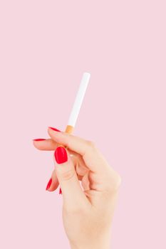 Female hand holding cigarette isolated on pink background. Feminine tobacco abuse.