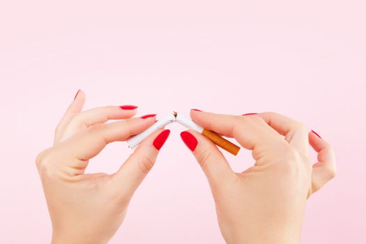 Female hands with red fingernails braking the last cigarette isolated on pink background. Quit smoking new year resolution.