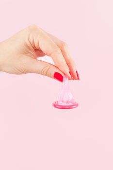 Female hand with red nails holding pink condom isolated on pink background. Safe sex and birth control concept.