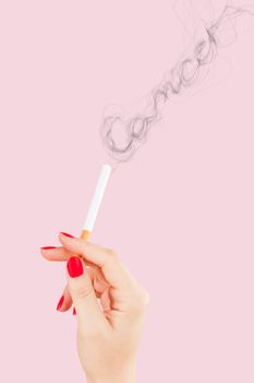 Female hand holding cigarette, smoke forming the word cancer isolated on pink background. Feminine tobacco abuse.