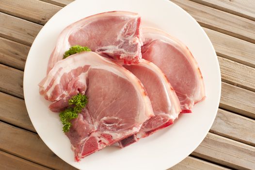 Four raw pork cutlets with fatty rind displayed ready for cooking on a plate on a wooden table, overhead view