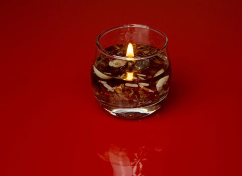 Burning decorative candle in a glass jar, filled with bits of shells and very small pebbles, isolated on a red background with a subtle reflection. Horizontal view.