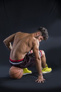 Young athletic shirtless man on dark background sitting on basketball ball, looking down, seen from the back