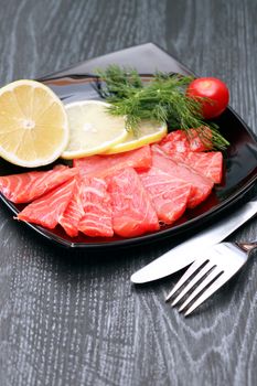 Slices of salted salmon with lemon on black plate near fork and knife