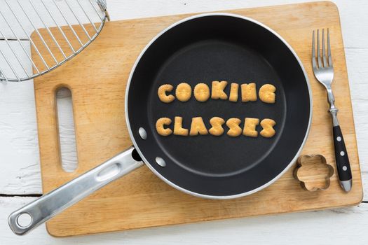 Top view of alphabet collage made of biscuits. Word COOKING CLASSES putting in black pan. Other kitchenware: fork, cookie cutter and chopping board putting on white wooden table, vintage style image.