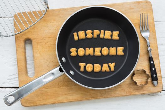 Top view of letter collage made of cookies. Quote INSPIRE SOMEONE TODAY putting in black pan. Other kitchen utensils: fork, cookie cutter and cutting board putting on white wooden table, vintage style image.