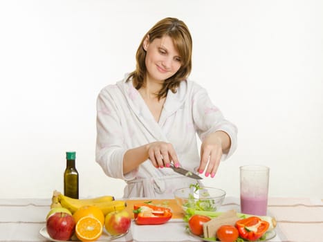 Young beautiful girl in a bathrobe prepares vegetarian salad from vegetables and greens isolated on white background