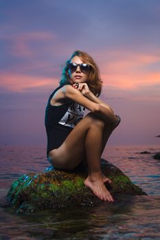 Beautiful young teen girl sitting on the stone in the sea fashion shoot at sunset beach