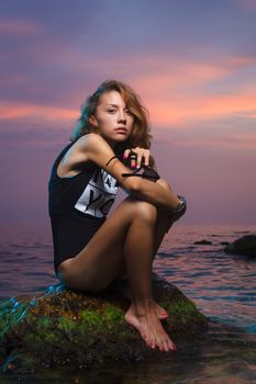 Beautiful young teen girl sitting on the stone in the sea fashion shoot at sunset beach
