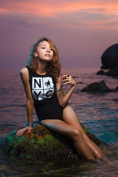 Beautiful young teen girl sitting on the stone in the sea fashion shoot at sunset beach