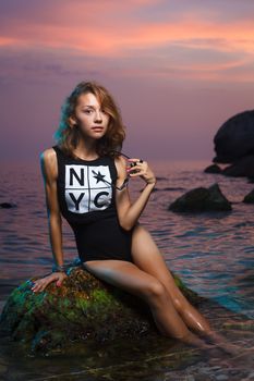 Beautiful young teen girl sitting on the stone in the sea fashion shoot at sunset beach