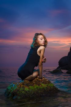 Beautiful young teen girl sitting on the stone in the sea fashion shoot at sunset beach