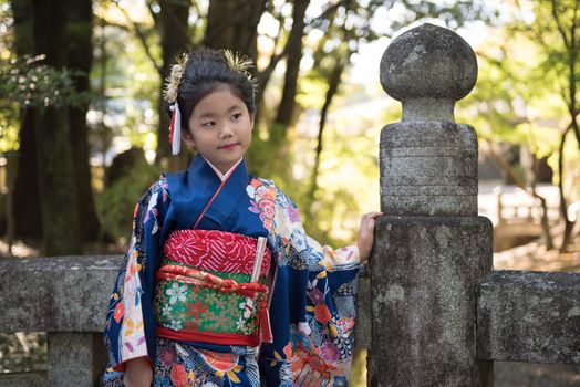 A young Japanese girl in a kimono outdoors on a stone bridge at a shrine.