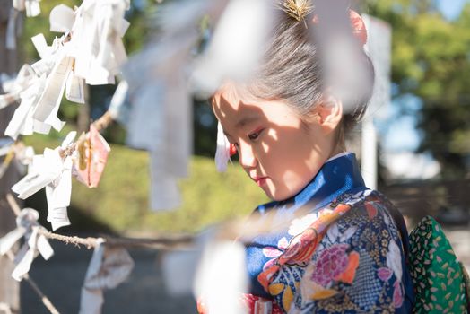 A young Japanese girl in a kimono outdoors at a shrine tying her fortune paper on a rope.