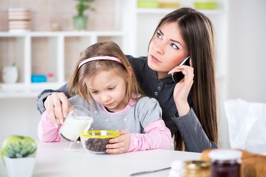 Overworked Business Woman and her little daughter in the morning. Overworked mother make phone calls before going to work. Preparing cereal with milk for her daughter and spilling milk next to bowl.