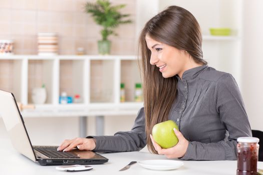 Businesswoman in the kitchen with apple reading mail on laptop before going to work.