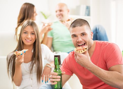 Close up of a young men smiling and eating pizza and drinking bear with her friends in the background.
