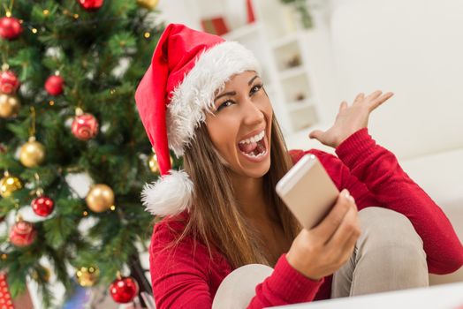 Cheerful young woman wearing Santa's hat is holding a smart phone at Christmas Time.