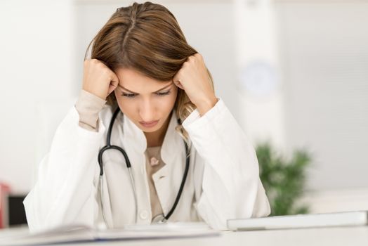 Frustration young female doctor sitting in her consulting room and looking at document.