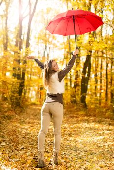 Beautiful cheerful young woman with red umbrella having fun in sunny forest in autumn colors.