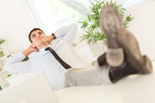 Young successful businessman in the office sitting on chair, hands behind  head with her feet up on a desk. He is relaxing and daydreaming.
