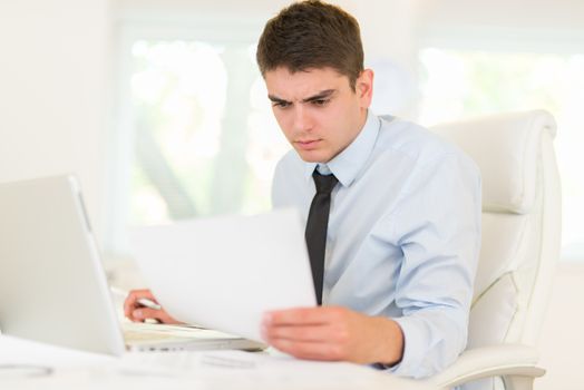 Young businessman sitting at an office desk in front of a laptop and looking at document.